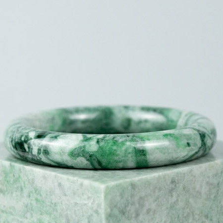 a single jade bangle atop a jade cube. the jade bangle is brecciated, mixing bright green with grey colors