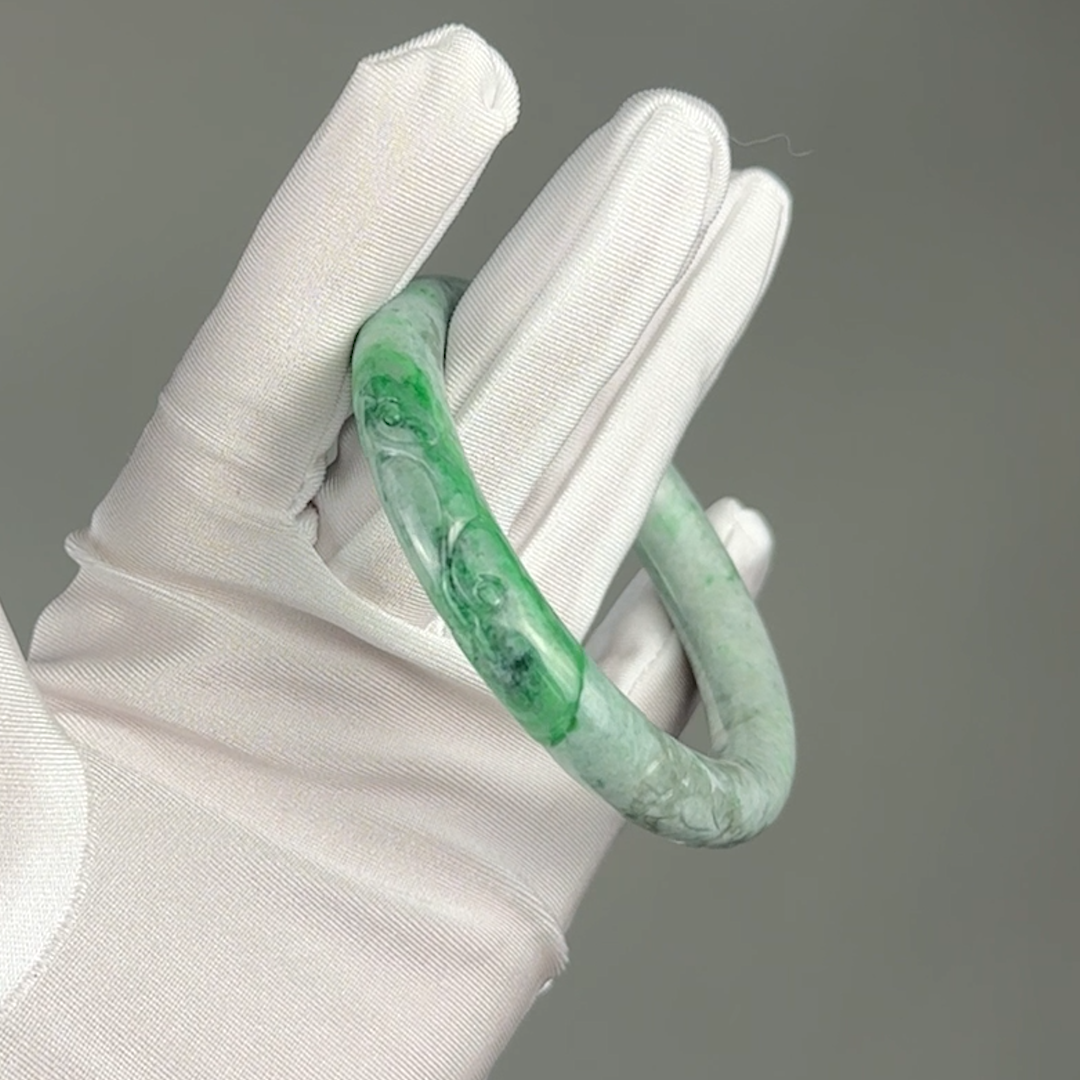 A green marbled jadeite bangle bracelet displayed on a white fabric glove against a solid gray background.