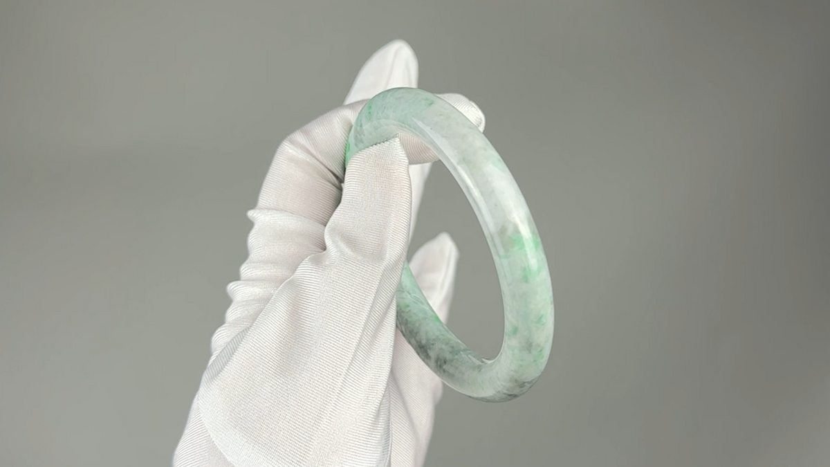 A light green and white marbled jadeite bangle held by a white gloved hand against a solid gray background.