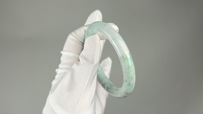 A light green and white marbled jadeite bangle held by a white gloved hand against a solid gray background.