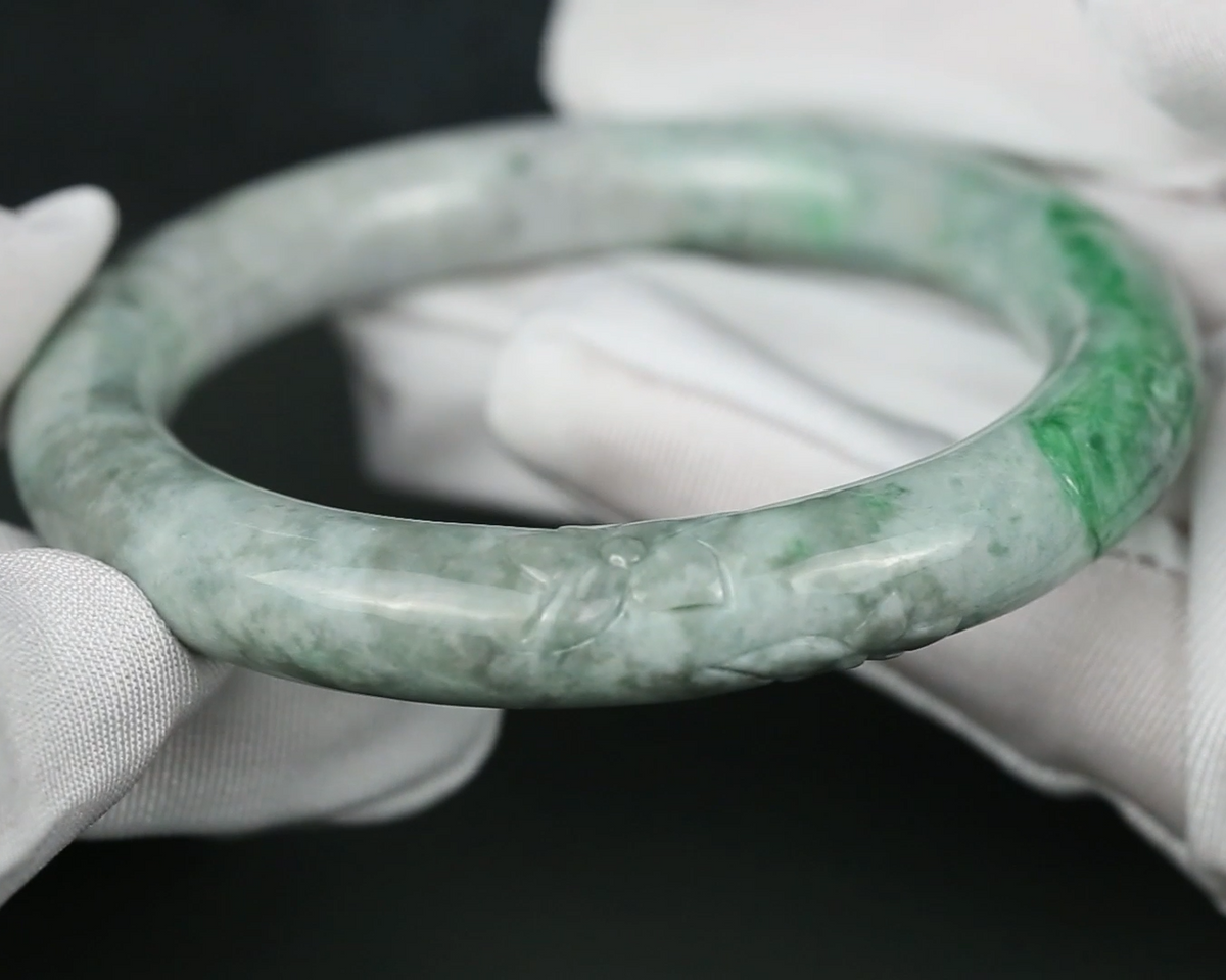 A carved jadeite bangle bracelet with green and white marbling, held by hands wearing white fabric gloves against a dark background.