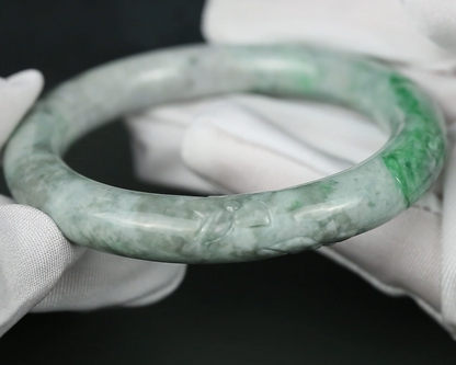 A carved jadeite bangle bracelet with green and white marbling, held by hands wearing white fabric gloves against a dark background.